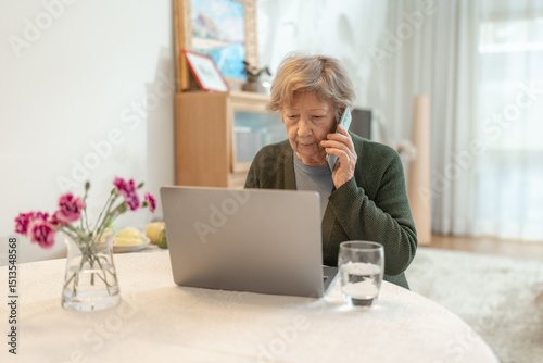 Senior woman using laptop at home while talking on the phone. Concept of technology, online shopping, digital literacy for seniors and active aging. Technology usage among seniors.