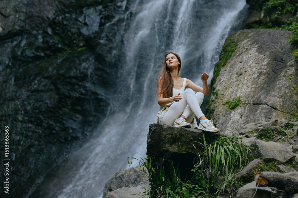 Obraz premium young woman in white clothes sitting on rock near waterfall surrounded by lush greenery in natural outdoor environment