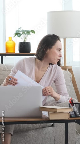 Focused woman working with paperwork and a laptop at home, while son uses a smartphone for leisure nearby, balancing tasks in a calm setting. Vertical video.