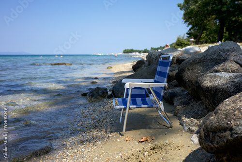 Fototapeta Naklejka Na Ścianę i Meble -  Empty blue striped beach chair sits on the rocky, pebbly shore of Stavros, Halkidiki, Greece, overlooking the calm Aegean Sea on a sunny summer day. Relaxing vacation scene by the water.	