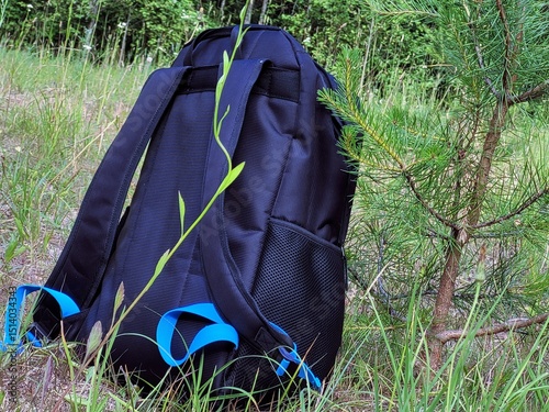 A close-up shot of a sleek black backpack with vibrant blue accents, resting on the ground in a lush, natural outdoor setting. The backpack features durable, textured fabric and functional mesh pocket