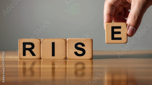 A hand placing a wooden block with the letter 'E' next to blocks spelling 'RIS', forming the word 'RISE'.