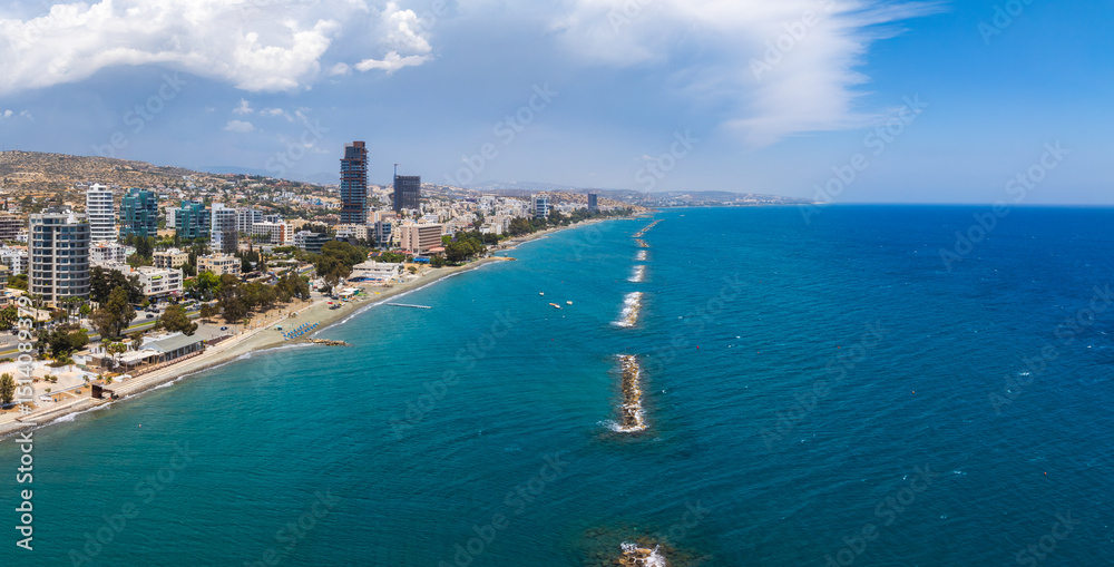 Fototapeta premium Aerial view of Limassol, Cyprus, showing modern high rises, sandy beaches, promenades, clear blue sea with breakwaters, and rolling hills under clouds.