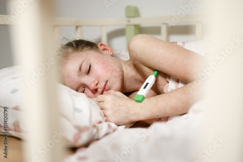 A young girl sleeping in a bed, appearing unwell. She is resting on a pillow with a thermometer in her hand, suggesting she may be sick. The scene is intimate and conveys a sense of care and concern.