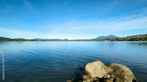 Serene landscape of Danau Diatas in West Sumatra, featuring a clear lake, rocky shore, green hills, and a bright blue sky.