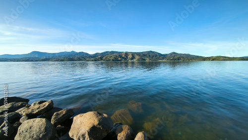 Serene landscape of Danau Diatas in West Sumatra, featuring a clear lake, rocky shore, green hills, and a bright blue sky.