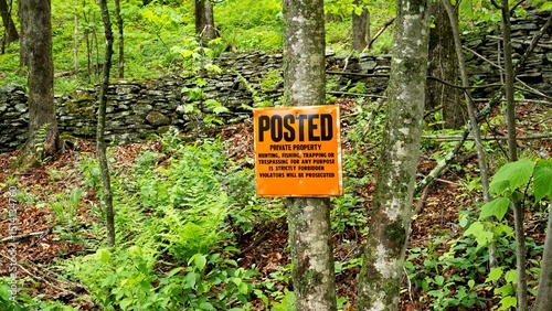 Orange POSTED sign in wooded area with trees and stone wall behind; Posted signs are often found in rural areas to indicate the land is private property and trespassing and other use is not allowed.