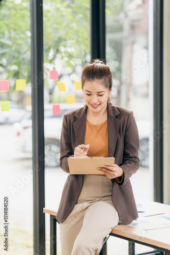 Free Download Happy Asian businesswoman writing on clipboard and smiling sitting at desk in office. Feeling happy and relaxed. Enjoying working in corporate workplace. - GFXLoad