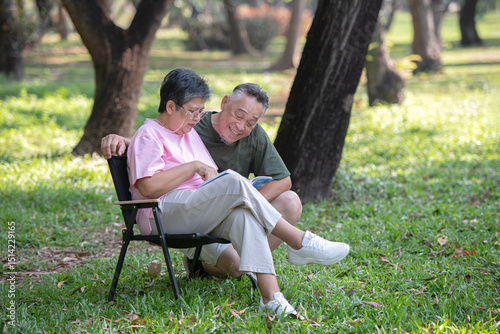 A retired couple is sitting on the lawn in a park reading a book and smiling happily.