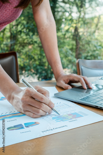 Close-up of a person reviewing financial reports with charts and graphs, using a laptop in a bright, modern office.