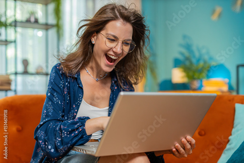 Surprised happy young woman in eyeglasses using laptop computer, receive good news message, mouth opened shocked by sudden victory celebrate lottery game jackpot win purchases online shopping at home.