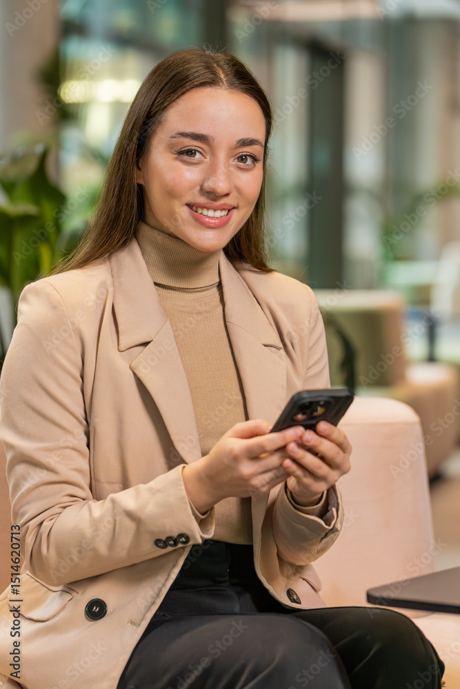 Fototapeta premium Caucasian young businesswoman girl using mobile phone texting share messages content working on smartphone social media applications online, relaxing in modern office lobby. Lady sitting in hotel hall
