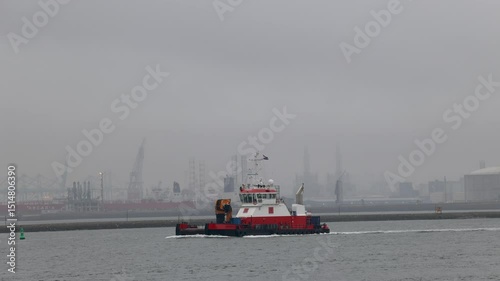Rotterdam, South Holland, Netherlands - 09.07.2024: A small red and white tugboat sails through a foggy industrial port, with cranes, ships, and cargo containers visible in the misty background.
