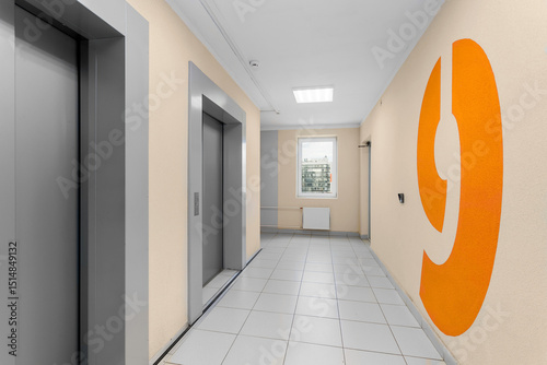 A spacious hall with elevators, a window at the end of the corridor, and a large orange number nine on a light wall.
