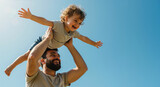 Happy bearded father lifting curly haired toddler son with arms spread against blue sky. Father's day celebration and family bonding activities