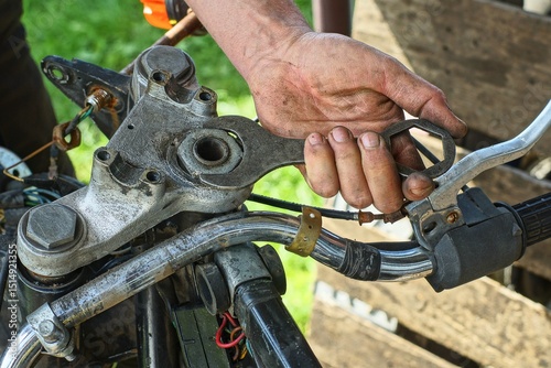 with one industrial iron wrench in his hand, a worker unscrews a nut in a telescopic gray fork on a motorcycle during the day on the street