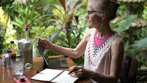 In a lush cafe, a mature woman with glasses engages with her tablet while jotting notes, embodying the spirit of a digital nomad on vacation.