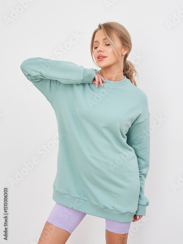 Blonde woman poses confidently, wearing an oversized teal sweatshirt and purple shorts, smiling with her right hand on her neck and left hand casually hanging. She stands against a light backdrop
