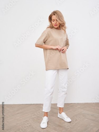 Woman wearing beige oversized shirt, white pants, and white sneakers, standing on light wooden floor. She has blonde hair and a relaxed pose, looking slightly down, hands together