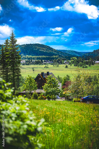 Country house surrounded by green fields, trees and mountains under a vivid blue sky with dramatic clouds.