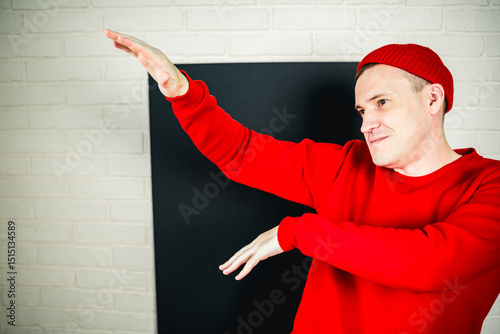 A man in a red hoodie and hat dances against a white brick wall