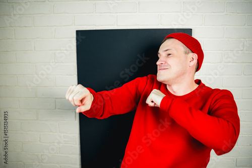A man in a red hoodie and hat dances against a white brick wall