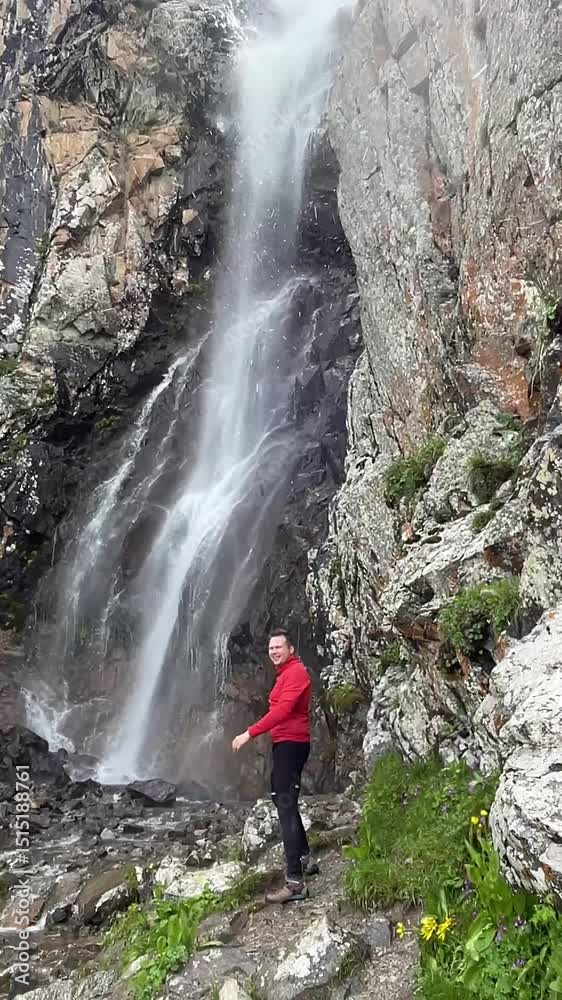 Kyrgyzstan, Ala Archa - 30.05.2025 - A mountaineer in red equipment stands at a waterfall, raises his hands, and water splashes into the camera. River in the mountains, wet rocks Kyrgyzstan, Ala Archa - 30.05.2025 - A mountaineer in red equipment stands at a waterfall, raises his hands, and water splashes into the camera. River in the mountains, wet rocks