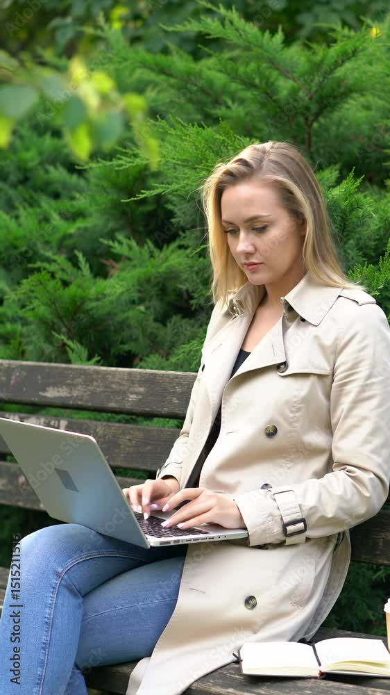Young caucasian woman in beige trench coat, blue jeans types on silver laptop on park bench with steaming coffee, open notebook, lush green bushes, soft dappled sunlight. Remote work in tranquil park