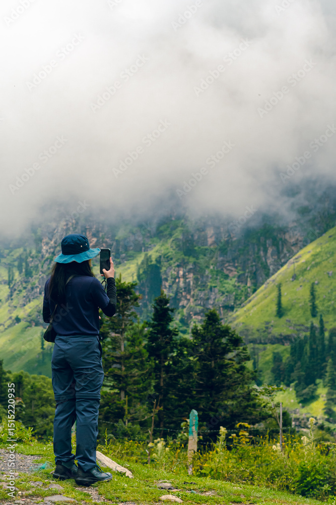 Obraz premium A woman Standing and taking pictures with a phone in a forest in India. She carries a shoulder bag and a blue bucket hat surrounded by lush green trees with foggy. The idea for an adventure activity.