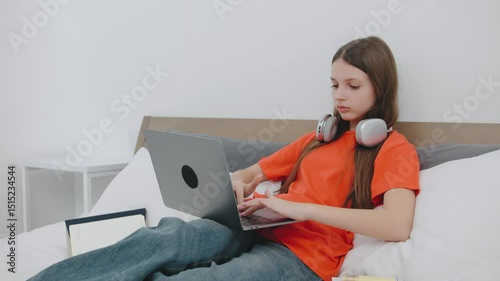 Teenage girl studies at home on a laptop while wearing headphones and comfortable clothing in a simple bedroom setting