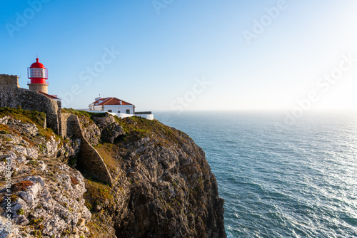 The iconic red and white lighthouse of Cabo da Roca stands atop a rugged cliff overlooking the expansive blue Atlantic Ocean under a clear sky
