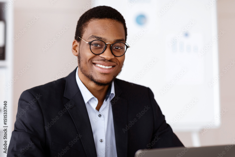 Businessman At Work. Portrait of happy african american young businessman in black suit sitting in front of laptop and looking at camera, sitting at desk in his modern office, working, copy space Businessman At Work. Portrait of happy african american young businessman in black suit sitting in front of laptop and looking at camera, sitting at desk in his modern office, working, copy space