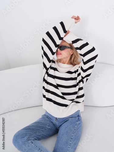Model poses cheerfully on a stylish couch while wearing a striped sweater and denim jeans