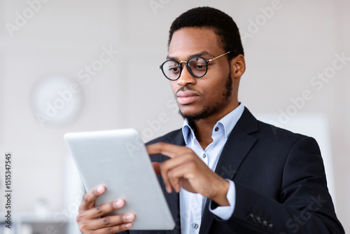 Closeup of serious black entrepreneur in glasses using modern digital tablet, checking trades and stocks online, office interior, copy space. Young african american manager holding digital pad