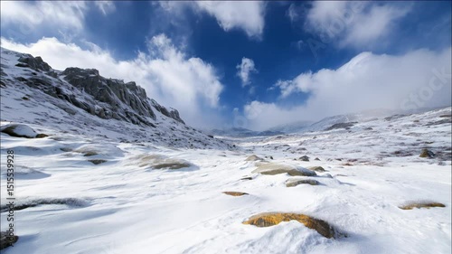 Snow-covered mountain slope with scattered rocks under bright blue sky and clouds in remote winter landscape