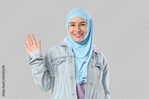 Happy young Muslim woman in hijab waving hand on grey background