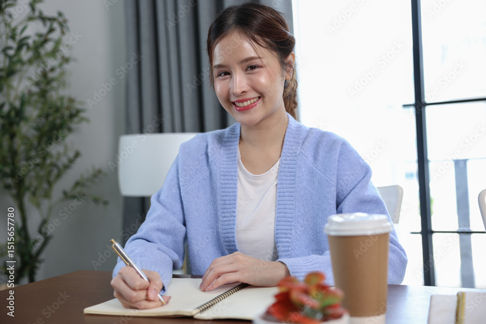 Asian cheerful woman in light blue cardigan is writing in notebook at wooden table, with coffee cup and small plant nearby, in bright and cozy indoor setting Asian cheerful woman in light blue cardigan is writing in notebook at wooden table, with coffee cup and small plant nearby, in bright and cozy indoor setting