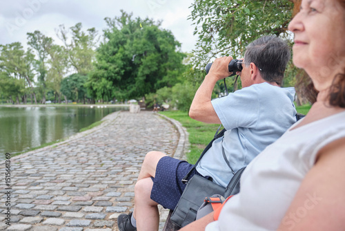 Senior Hispanic couple relaxing together in a park. In the foreground the woman looks at the lake in front of her, behind her the man uses binoculars to spot birds. Composition with selective focus.
