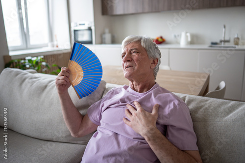 Distressed older man struggling with unbearable heat, using fan to relieve suffocation and nausea. Heat stress impacting health, lack of air, cardiovascular pressure, unwell in sweltering home 