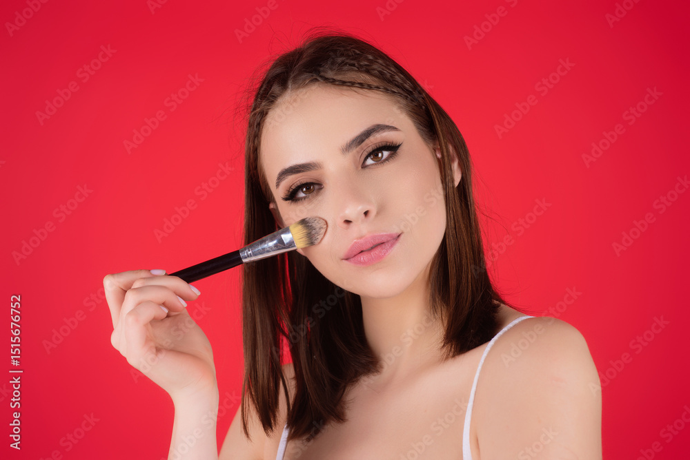 Obraz premium Beautiful young woman applying makeup on studio background. Pretty girl holding makeup brushes and make up on face with cosmetics. Facial beauty. Perfect skin, natural make up.