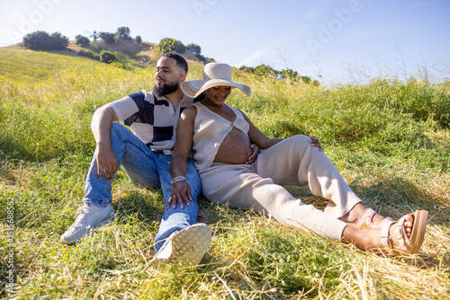 An African American couple relaxes in nature, joyfully celebrating their pregnancy and love for each other amidst a vibrant summer landscape filled with greenery and warm sunshine