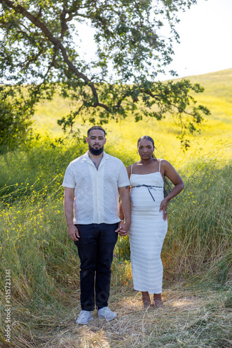 A happy and expectant couple enjoys the beautiful outdoors on a summer day, standing together in a sunlit area while the woman proudly displays her baby bump, surrounded by natures vibrant beauty