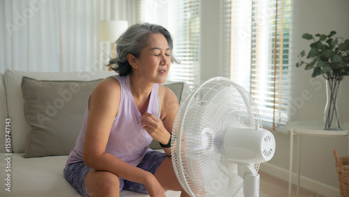 Asia adult people middle age old woman sit at sofa home feel hot flash fever use fan waving hand blowing wind face elder health care summer heat aging issue. Sad pain sweat tired stroke suffer stress.