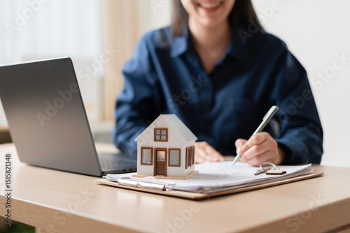 homebuyer signing property purchase agreement at desk with laptop and miniature house model, finalizing mortgage and closing transaction