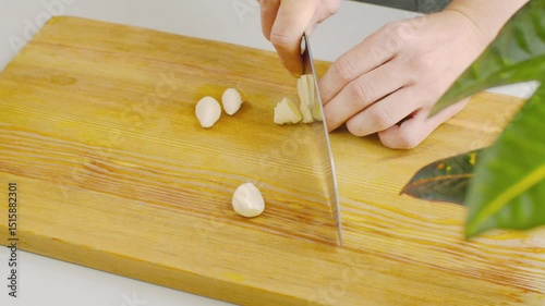A person slices a garlic clove into thin pieces on a cutting board. Close-up of the knife cutting through the white aromatic clove in a bright kitchen with natural light.