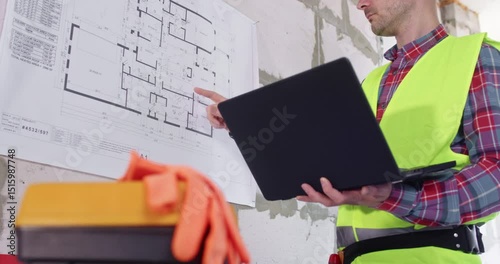 Specialist studies floor plan fixed on wall holding computer in hands. Professional uses modern laptop to perform special calculations and analyze building layout
