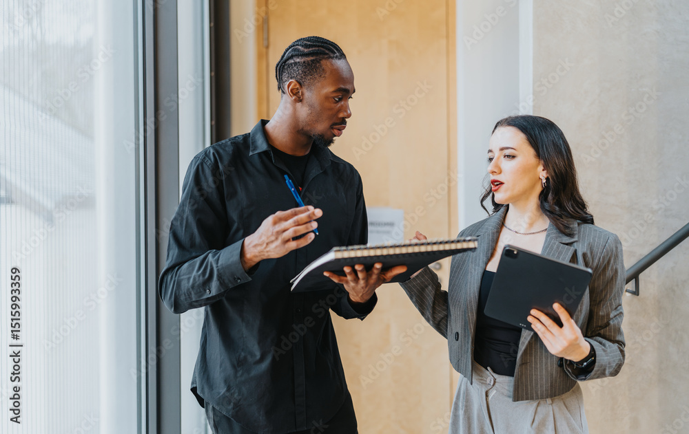 Fototapeta premium Two diverse people engaged in a vibrant discussion about work, highlighting teamwork and modern office collaboration using tools like notebooks and tablets in an innovative workplace.