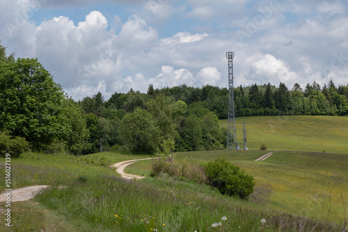 Telecommunication tower in a rural area improving mobile signal coverage, surrounded by fields, forest, and a winding dirt road under a partly cloudy sky.