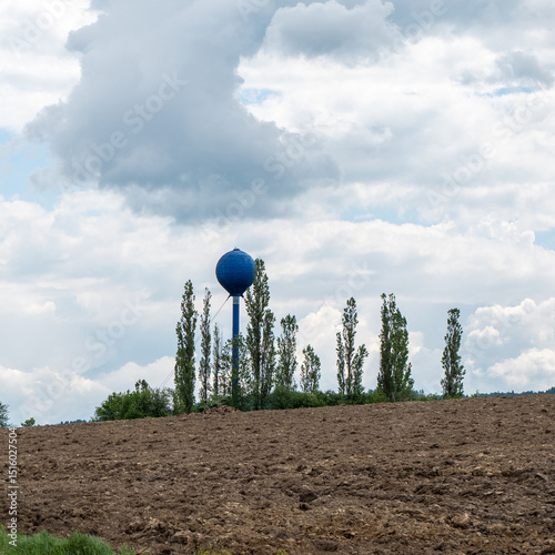 Blue elevated water tank used for agricultural irrigation, standing behind plowed farmland with tall trees and a cloudy sky in the background.