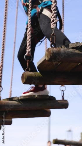 View of high ropes course, kids of climbing in amusement acitivity rope park, passing obstacles and zip line on heights, children teenagers in equipment gear between the trees on heights, summer day